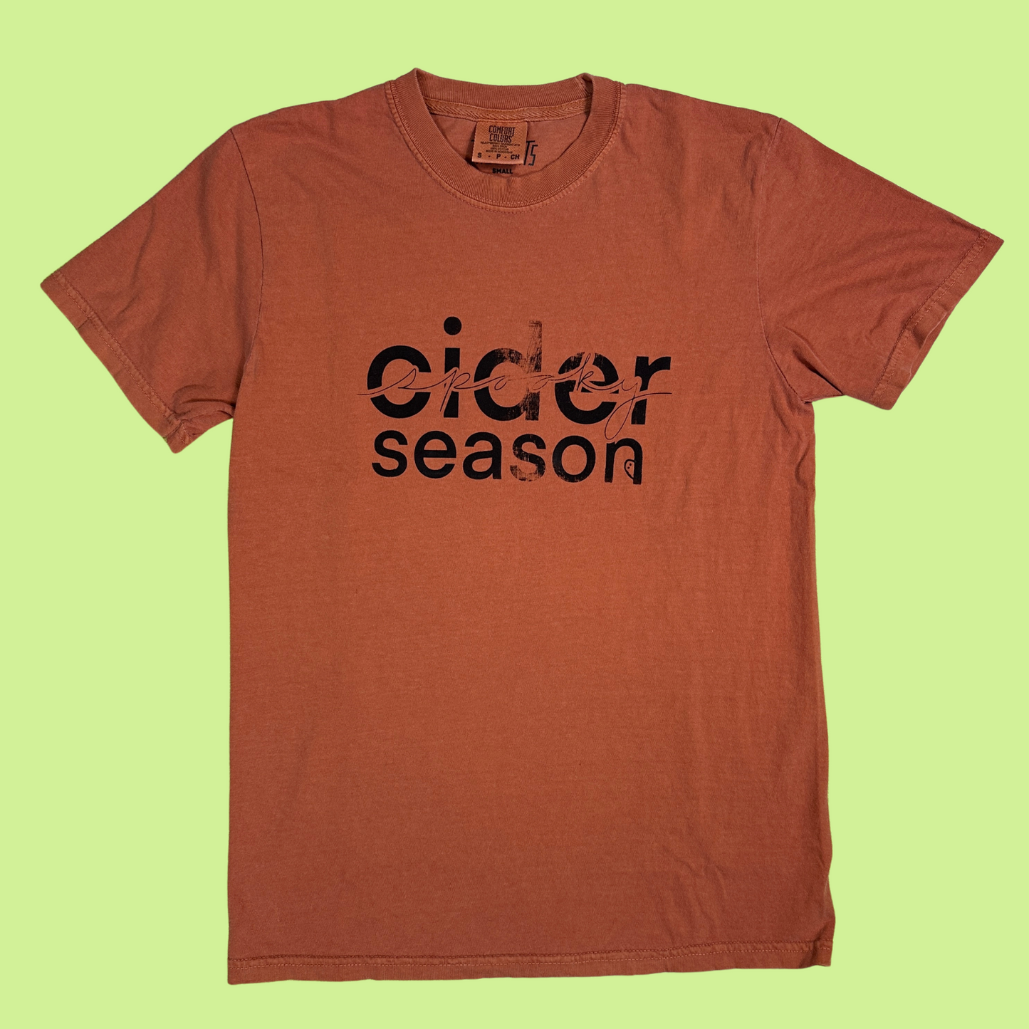 Rust/orange comfort colors unisex tshirt with "cider season" in bold text with a little distressing in the design with "spooky" in cursive running through "cider". Has a little ghost peaking out of the "n" in "season".