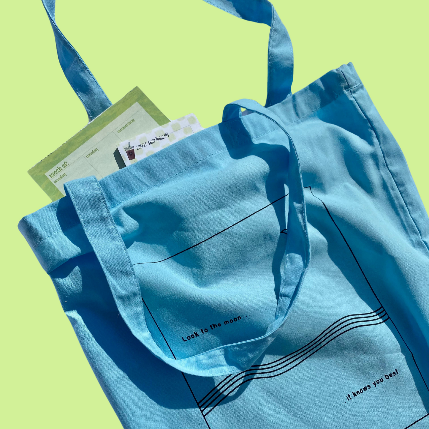 Light Blue Lightweight Tote Bag with matching fabric handles. Rectangle design with wave detail like the ocean with a moon in the sky and the saying "Look to the moon, it knows you best" graphic is a black outline/text on the light blue tote bag. In this photo, the tote bag is used to carry other Inner Thoughts products like a weekly "to do" notepad and "coffeeshop thoughts" sticky notes. Not ideal for heavy things like books or laptop, but perfect for a farmer's market or everyday essentials.