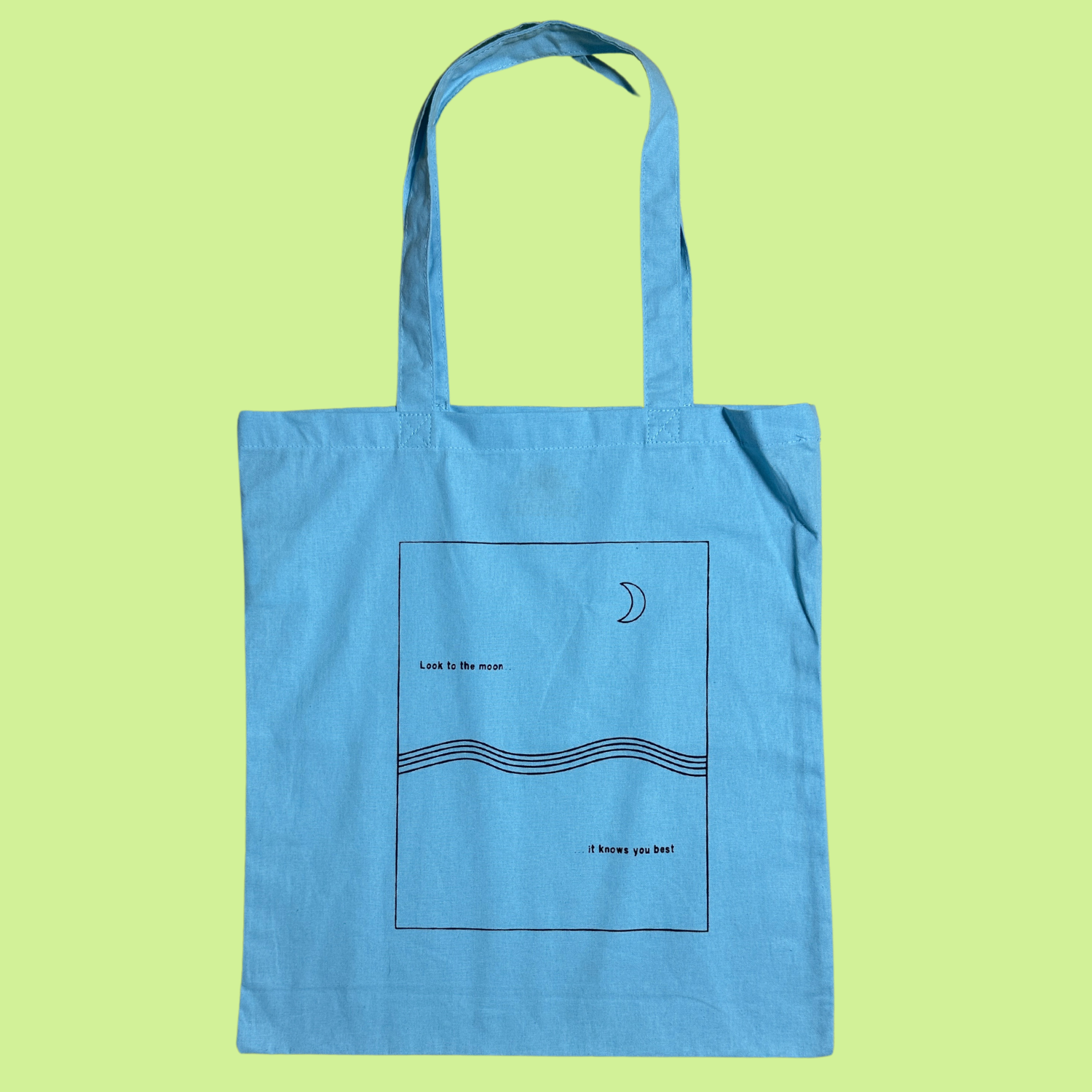 Light Blue Lightweight Tote Bag with matching fabric handles. Rectangle design with wave detail like the ocean with a moon in the sky and the saying "Look to the moon, it knows you best" graphic is a black outline/text on the light blue tote bag. Not ideal for heavy things like books or laptop, but perfect for a farmer's market or everyday essentials.